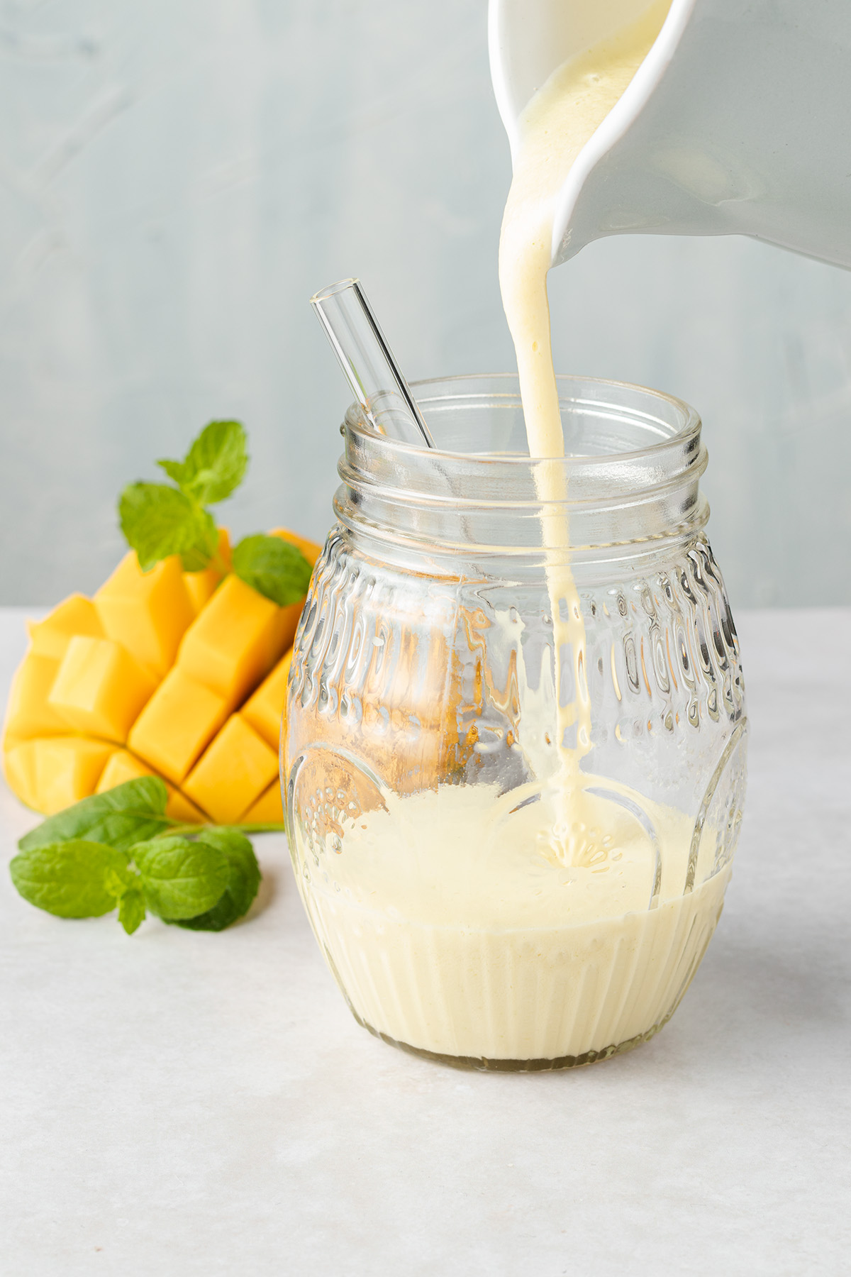 Ananas Smoothie wird in eine Glas eingegossen, im Hintergrund sieht man eine Mango und Minzbl&auml;tter