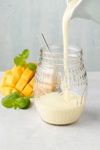 Ananas Smoothie wird in eine Glas eingegossen, im Hintergrund sieht man eine Mango und Minzbl&auml;tter