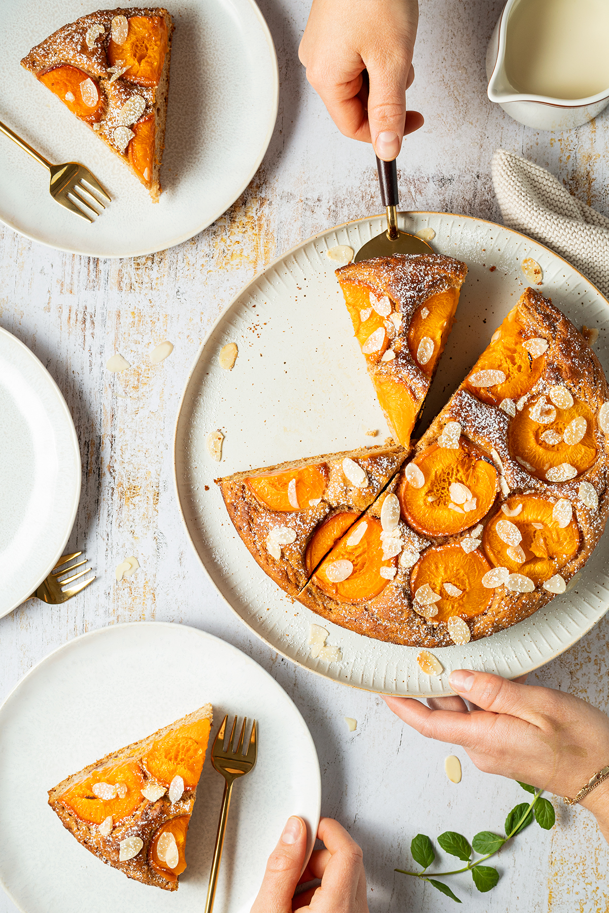 Ein wei&szlig;er Holztisch mit dem Aprikosenkuchen auf einem gro&szlig;en Tortenteller, zwei St&uuml;cke des Kuchens liegen bereits auf wei&szlig;en Kuchentellern, ein weiteres wird gerade auf einen dritten Teller gehoben.