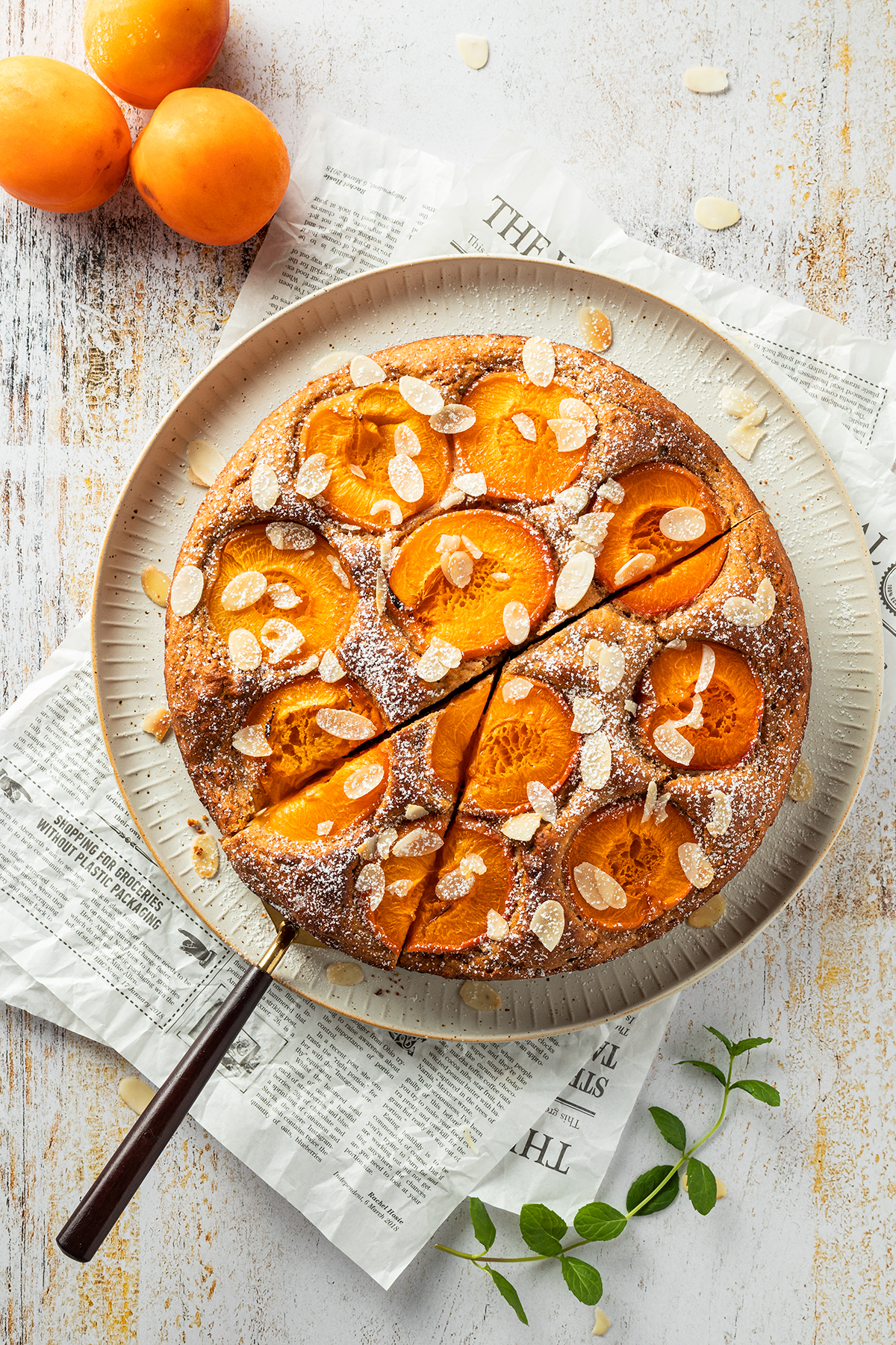 Aprikosenkuchen mit Mandelbl&auml;ttchen und Puderzucker oben drauf, daneben liegen drei Aprikosen un dein Zweig frische Minze