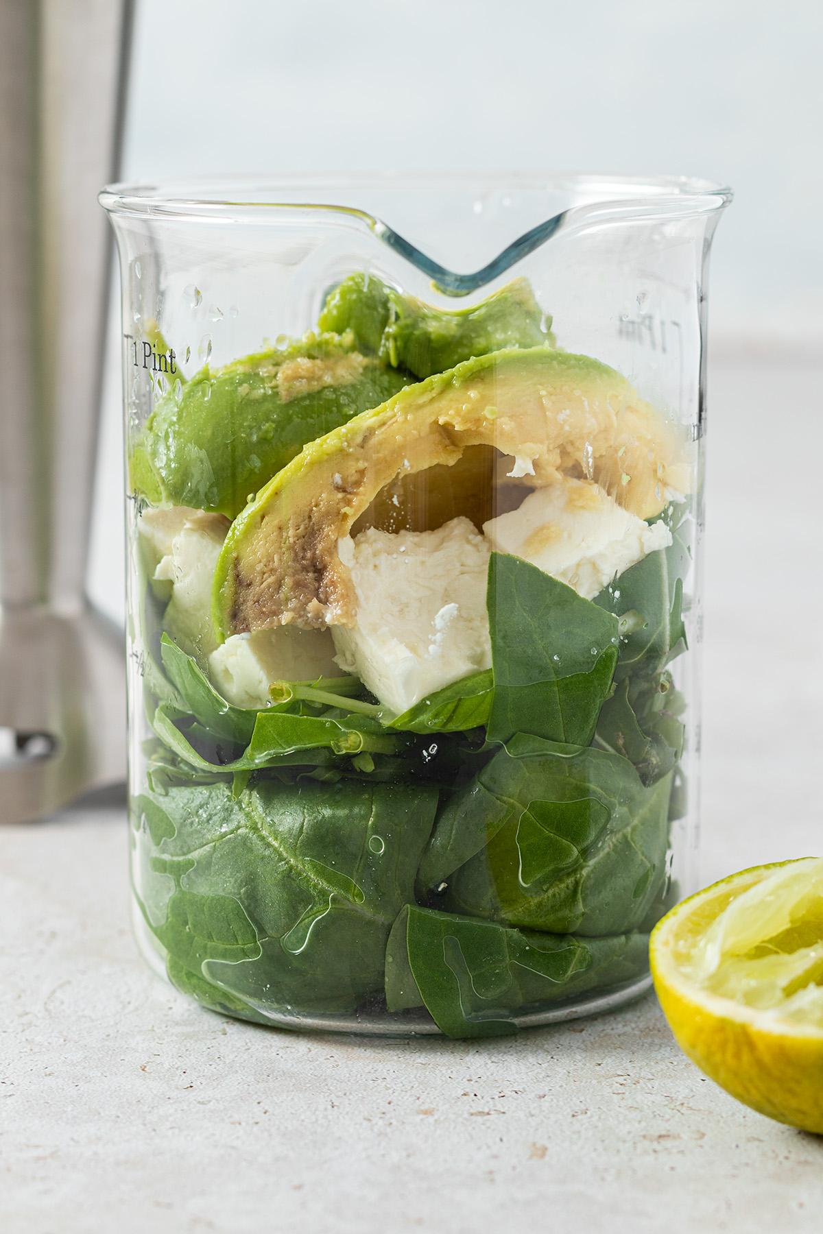 Spinat, Feta und Avocado in einem Messbecher, im Vordergrund eine ausgepresste Limette, im Hintergrund ein P&uuml;rierstab