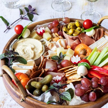 Eine reichlich gef&uuml;llte Brotzeitplatte mit K&auml;se, Schinken, Dips, Gem&uuml;se, Obst und Cracker. Im Hintergrund stehen Wein und Wassergl&auml;ser und noch mehr Cracker.