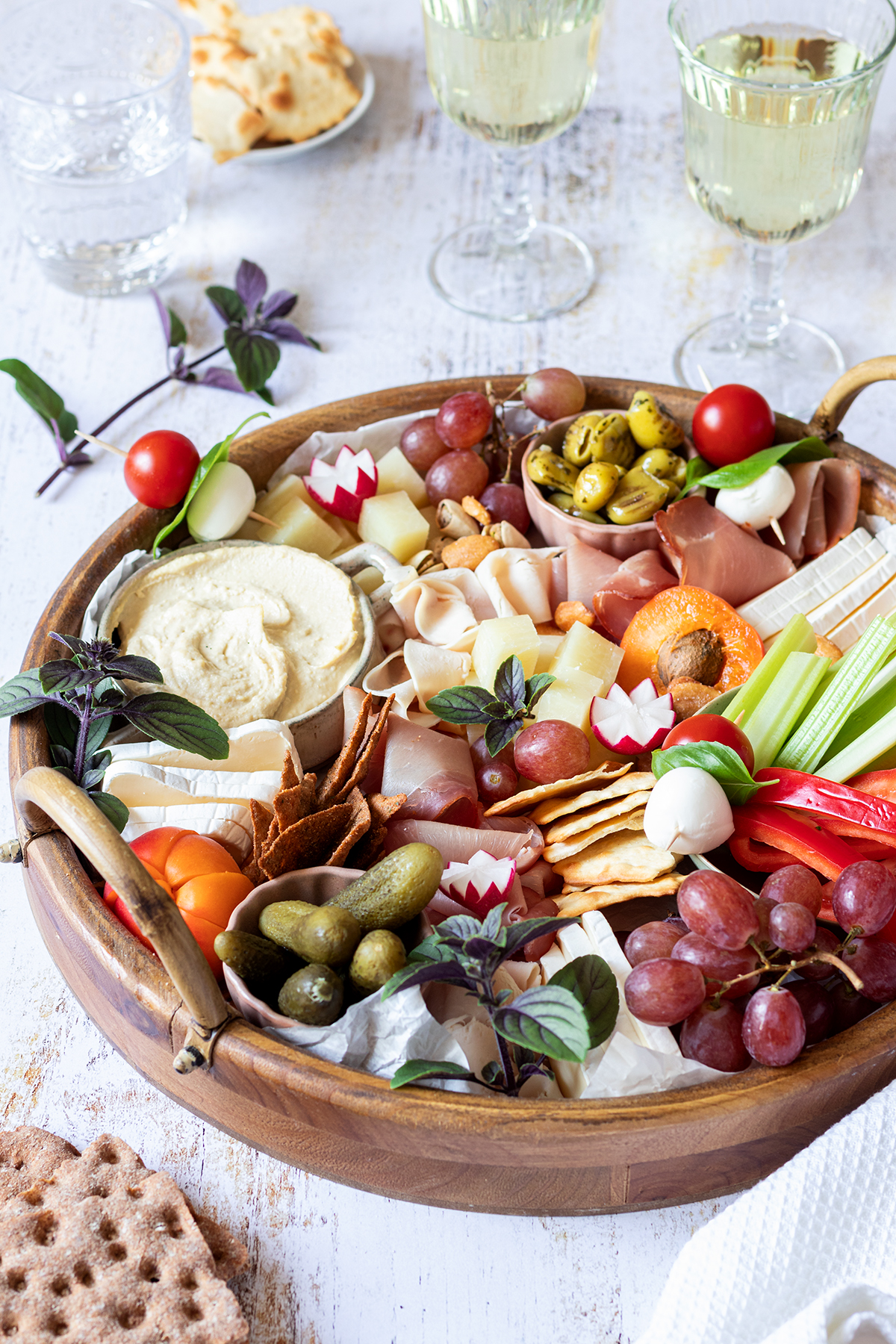 Eine reichlich gef&uuml;llte Brotzeitplatte mit K&auml;se, Schinken, Dips, Gem&uuml;se, Obst und Cracker. Im Hintergrund stehen Wein und Wassergl&auml;ser und noch mehr Cracker.