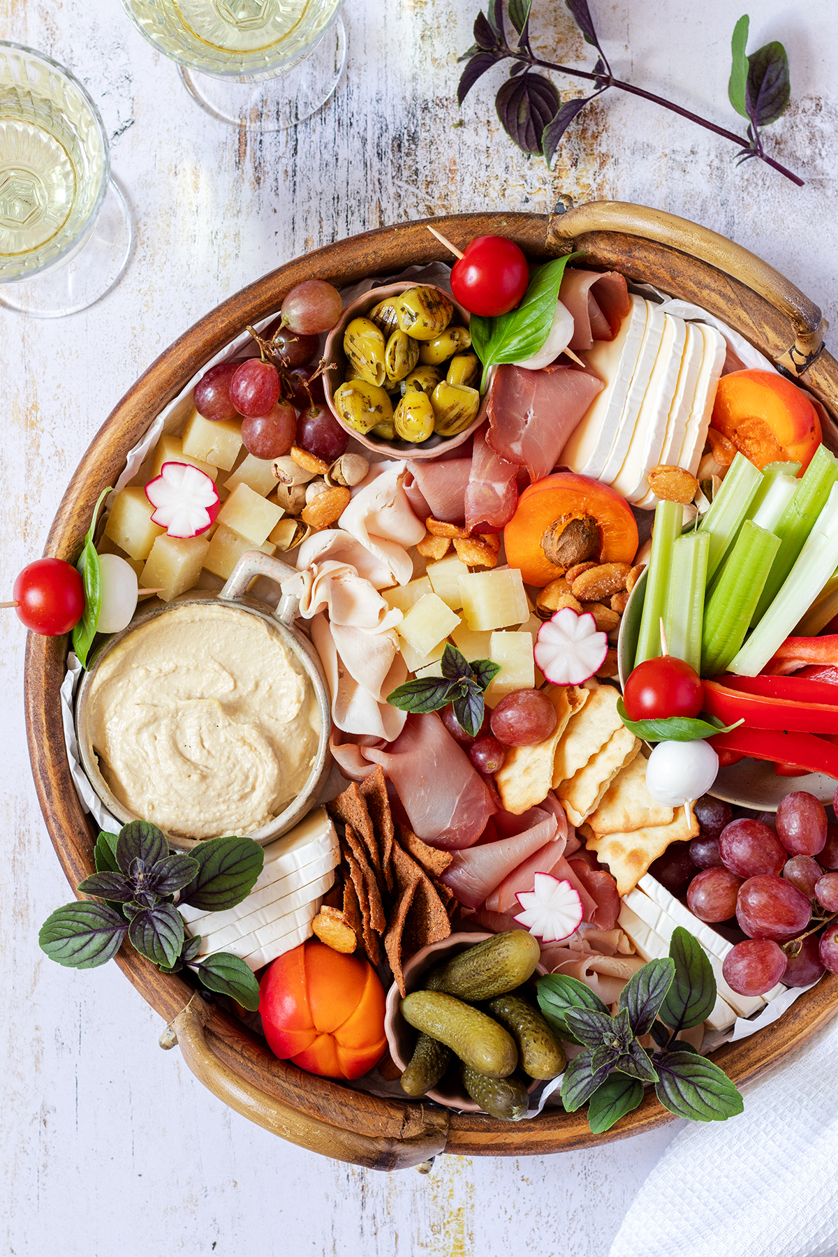 Eine reichlich gef&uuml;llte Brotzeitplatte mit K&auml;se, Schinken, Dips, Gem&uuml;se, Obst und Cracker. Im Bildrand stehen Wein- und Wassergl&auml;ser.