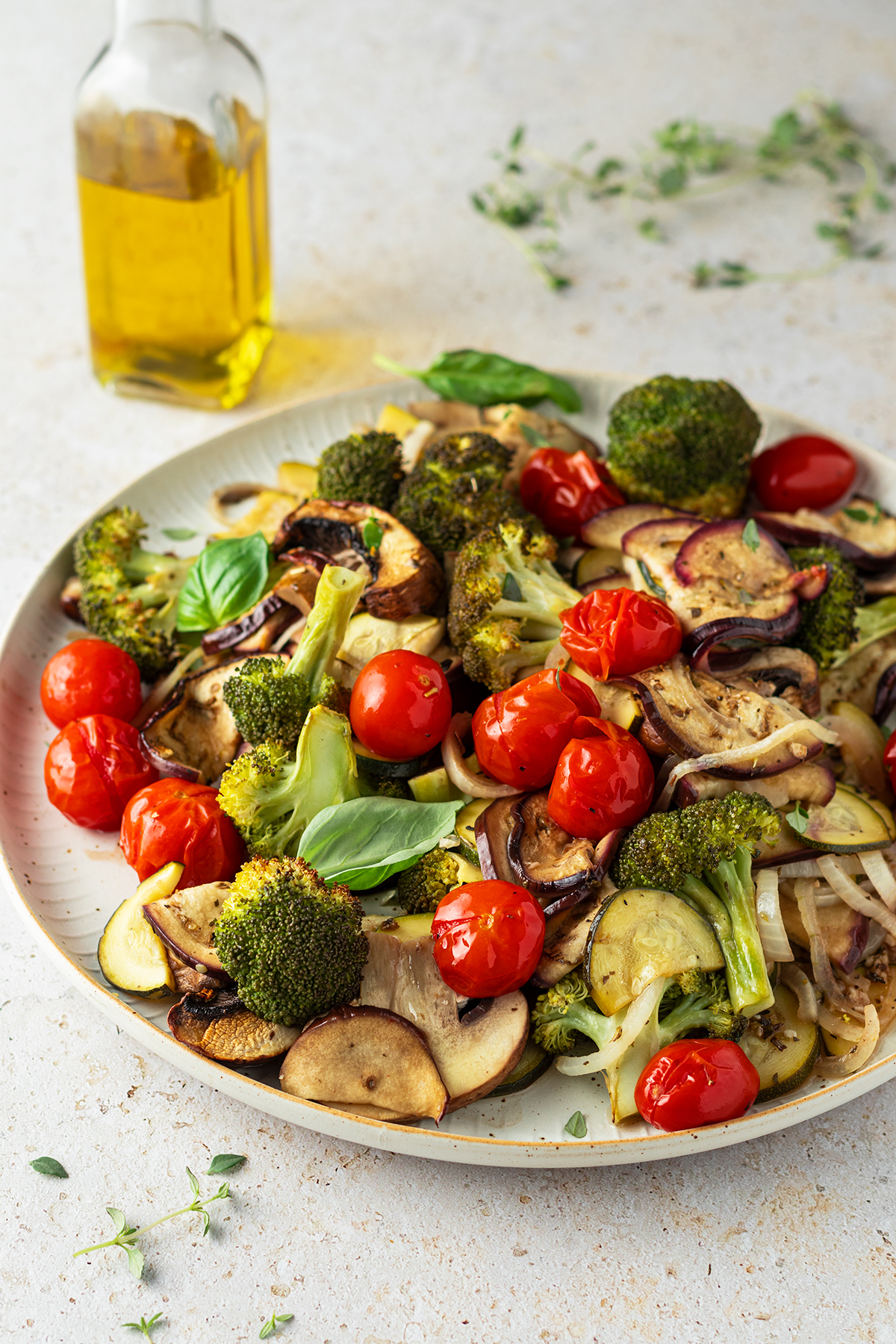 Buntes Ofengem&uuml;se vom Blech mit Tomaten, Brokkoli, Aubergine und Zucchini serviert auf einem gro&szlig;en, wei&szlig;en Teller. Im Hintergrund eine Flasche Oliven&ouml;l und frische Kr&auml;uter