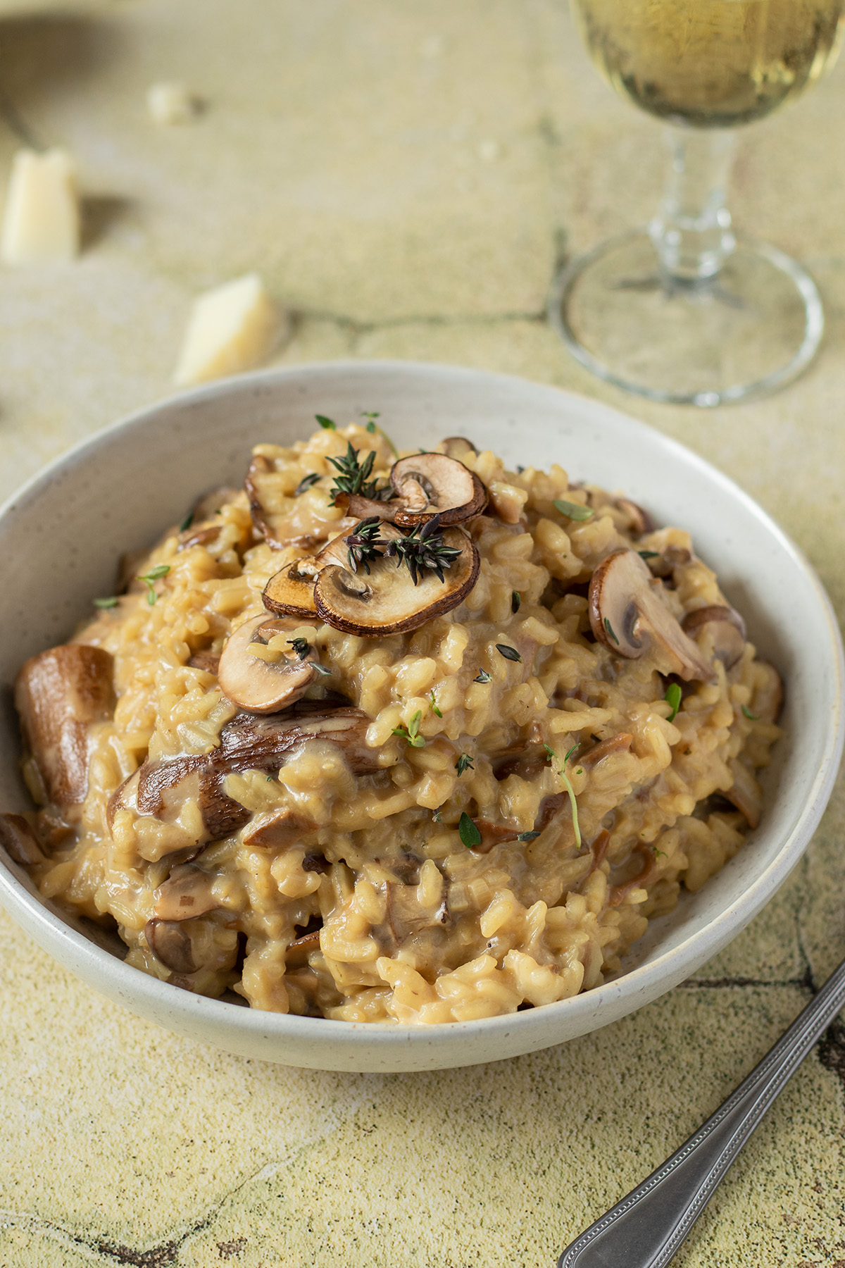 Teller mit cremigen Steinpilzrisotto auf gr&uuml;nen Flie&szlig;en, Parmesan und ein Weinglas im Hintergrund.