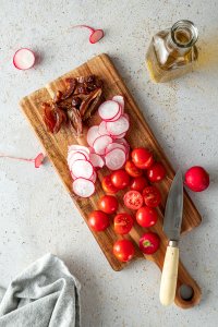 Halbierte Cherrytomaten, d&uuml;nne Radieschen Scheiben und Datteln auf einem Schneidebrett, daneben ein Schneidemesser und eine Flasche Oliven&ouml;l.