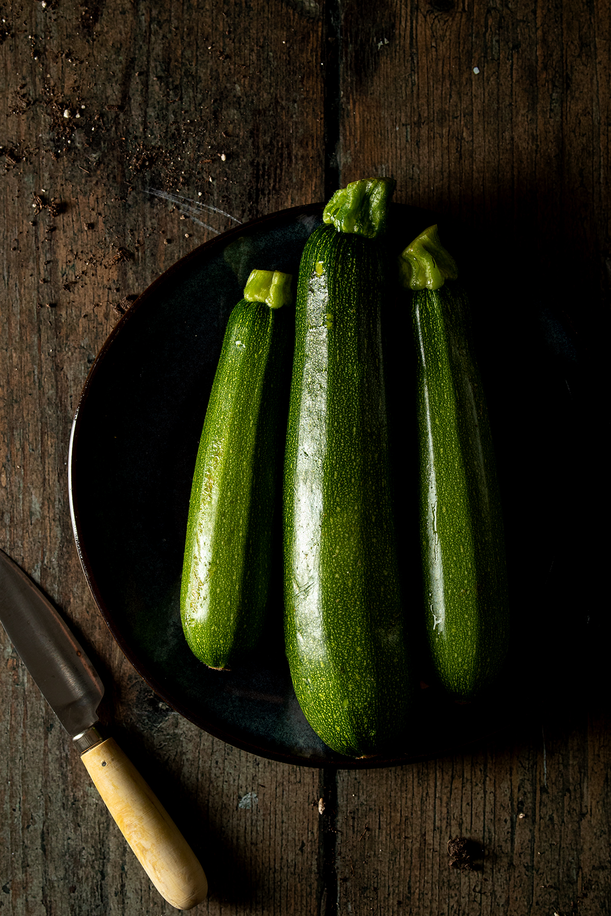 Drei Zucchini auf einem dunklen Teller, der auf einem Holztisch steht.
