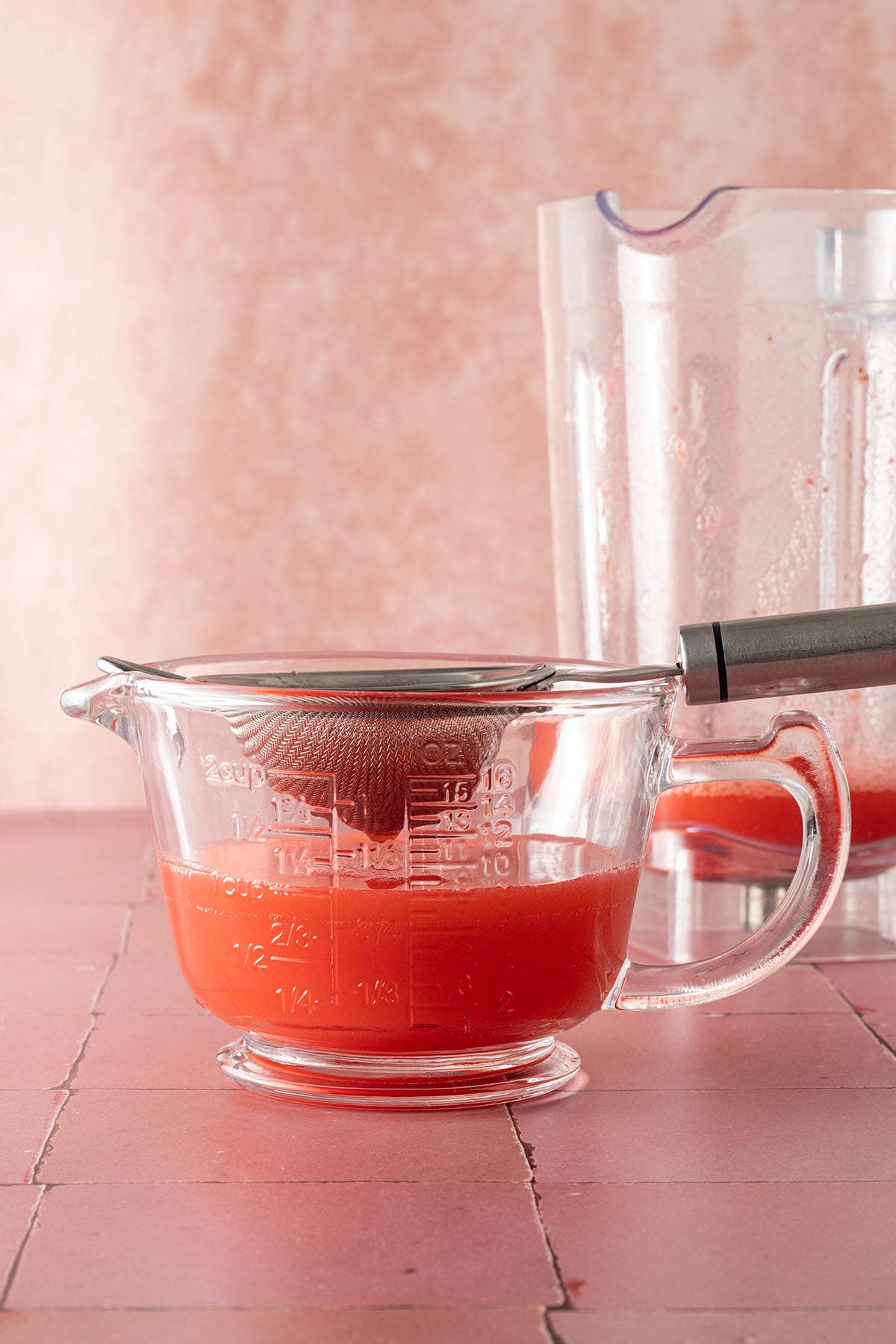 Erdbeer-Agua-Fresca wird durch ein feines Sieb in einen Messbecher gegossen, im Hintergrund der Mixerbeh&auml;lter.