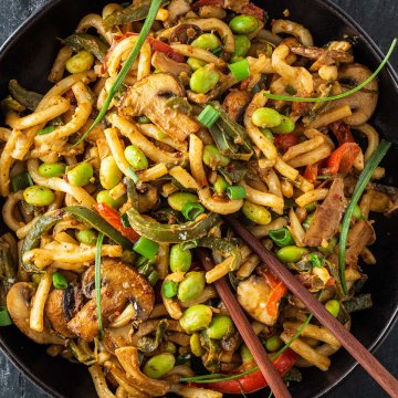 Eine Portion Udon Nudeln mit Gem&uuml;se, einer cremigen Erdnusssauce und Edamame Bohnen auf einem Teller mit Essst&auml;bchen.