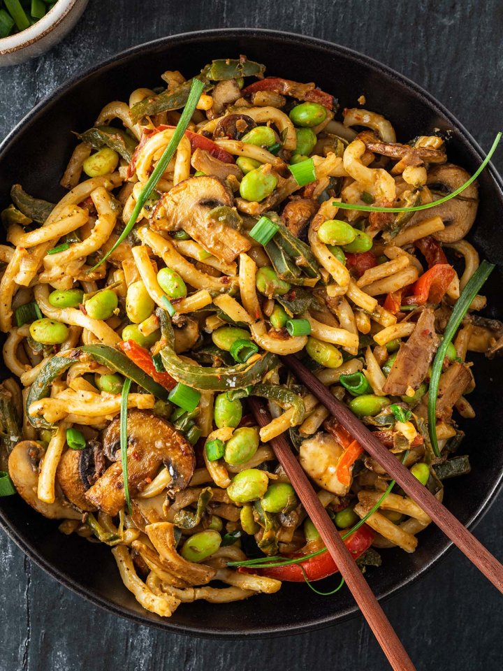 Eine Portion Udon Nudeln mit Gem&uuml;se, einer cremigen Erdnusssauce und Edamame Bohnen auf einem Teller mit Essst&auml;bchen.