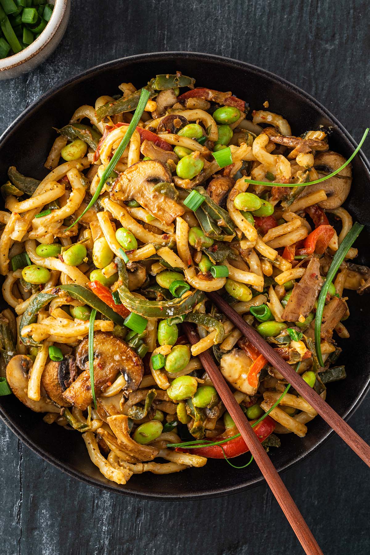 Eine Portion Udon Nudeln mit Gem&uuml;se, einer cremigen Erdnusssauce und Edamame Bohnen auf einem Teller mit Essst&auml;bchen.
