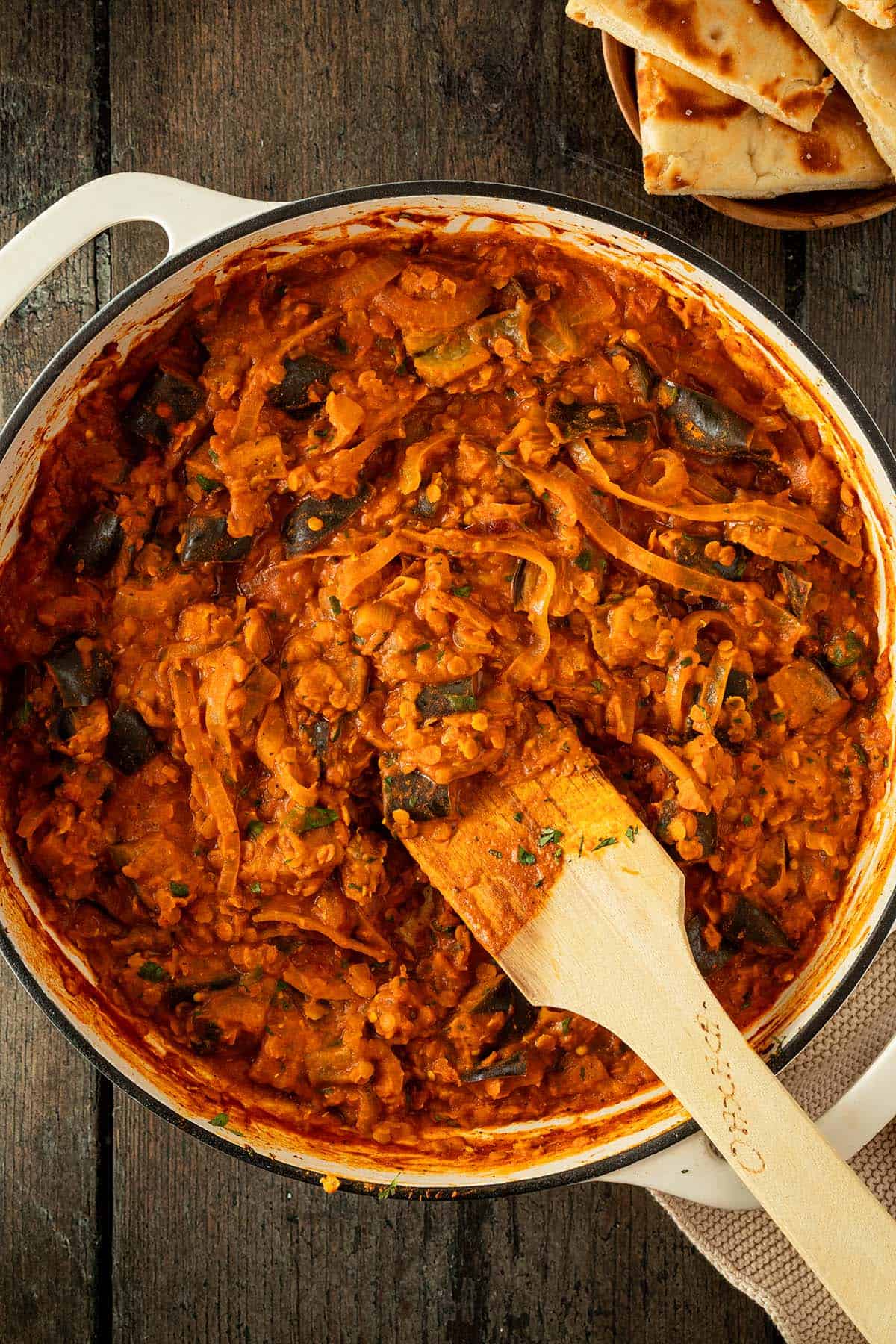 Auberginen Curry mit Linsen und Zwiebeln in einer cremigen Tomatensauce in einer gro&szlig;en Pfanne auf einem rustikalen Holzuntergrund, im Bildrand Naan.
