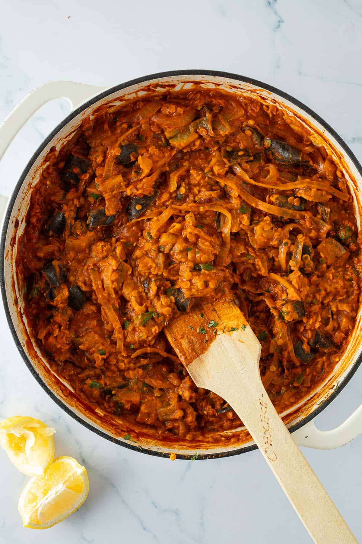 Fertig gegartes Auberginen Linsen Curry aus dem Ofen in einer gro&szlig;en Pfanne, daneben zwei Zitronenspalten.