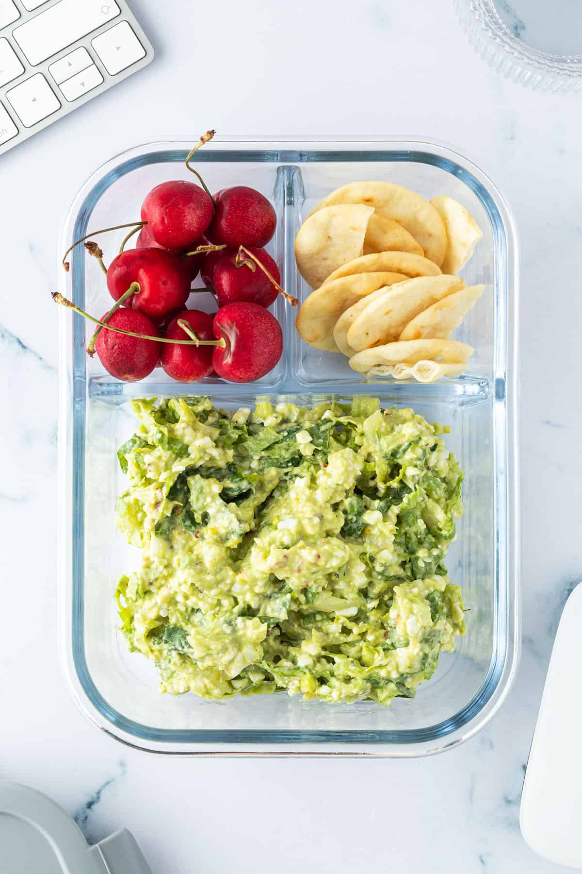 Avocado Eier Salat in einer Lunchbox mit Crackern und Kirschen, Scheibtisch-Gegenst&auml;nde liegen im Bildrand.