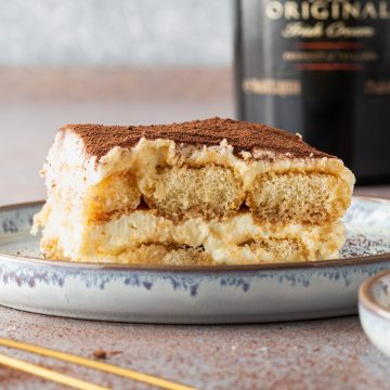 Ein St&uuml;ck Tiramisu auf einem Teller mit einer Flache Bailey im Hintergrund.