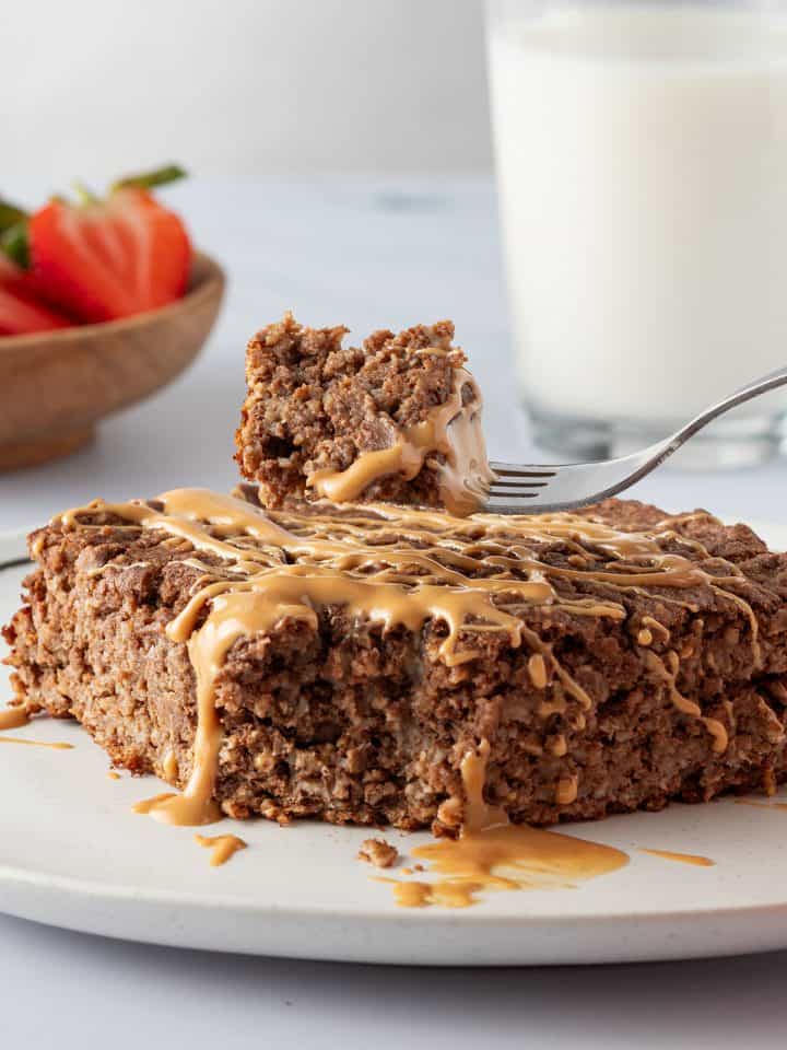 Ein Bissen vom Brownie Baked Oatmeal auf einer Gabel, Milch und Erdbeeren im Hintergrund.