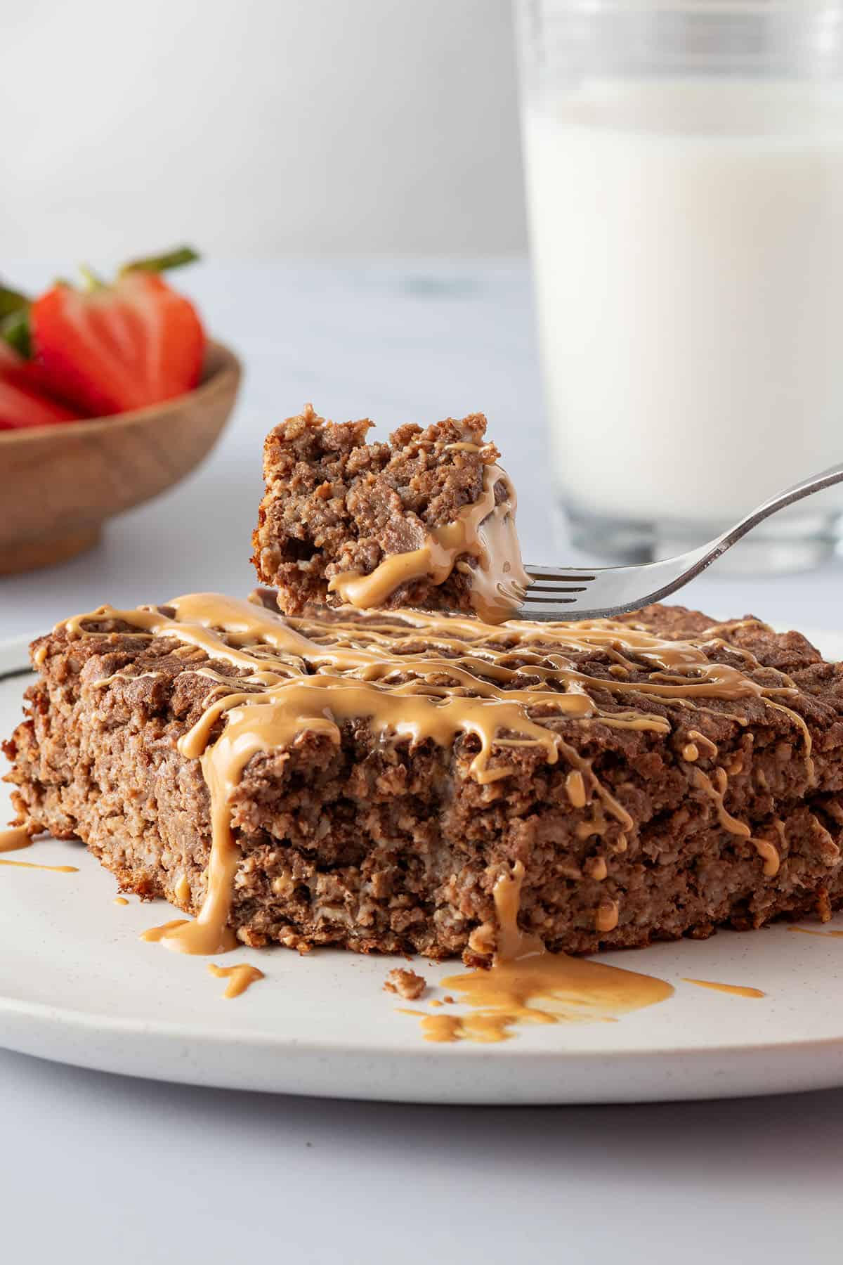 Ein Bissen vom Brownie Baked Oatmeal auf einer Gabel, Milch und Erdbeeren im Hintergrund.