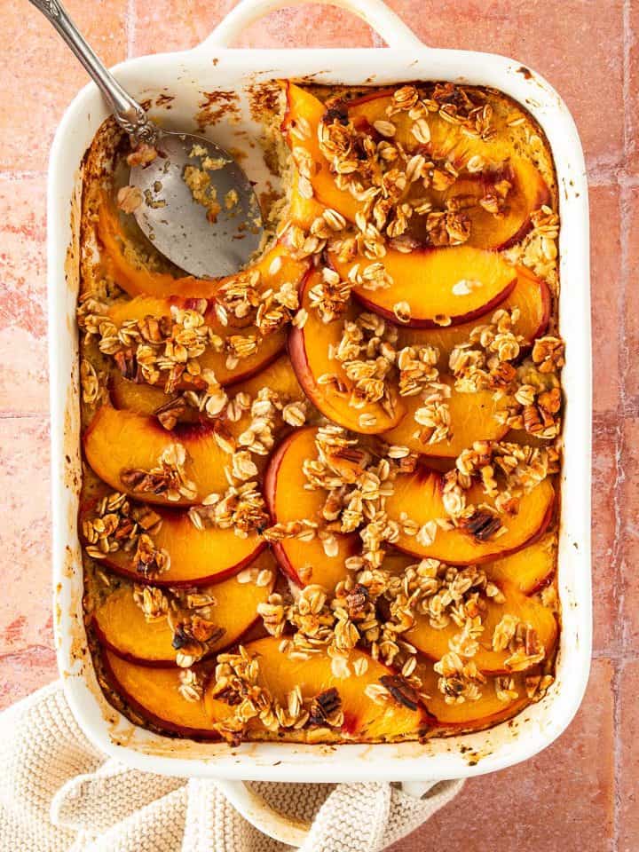 Pfirsich Baked Oatmeal in einer Auflaufform auf einem roten Fliesen Untergrund. Ein L&ouml;ffel eine Portion des Haferflockenauflaufs herausgenommen.