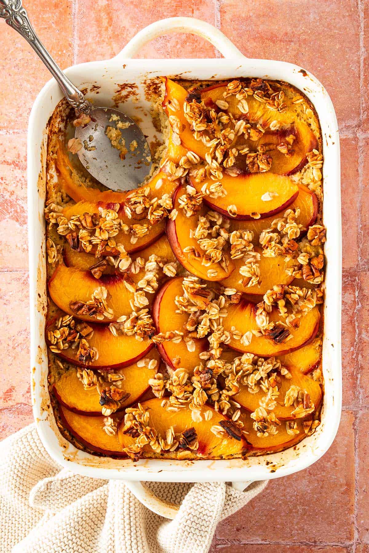 Pfirsich Baked Oatmeal in einer Auflaufform auf einem roten Fliesen Untergrund. Ein L&ouml;ffel eine Portion des Haferflockenauflaufs herausgenommen.