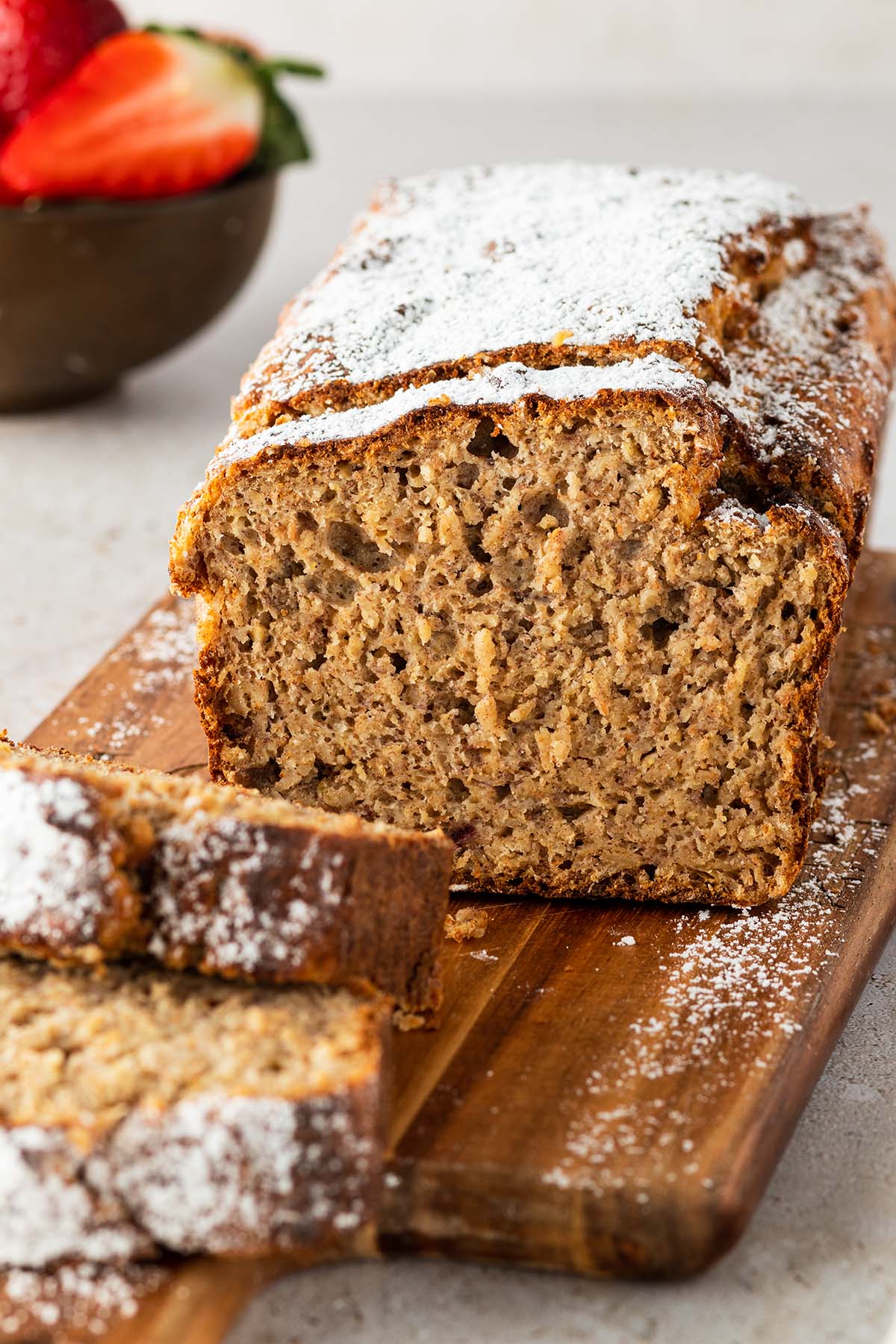 Bananenbrot mit Haferflocken und Puderzucker serviert mit einer Sch&uuml;ssel Beeren im Hintergrund.
