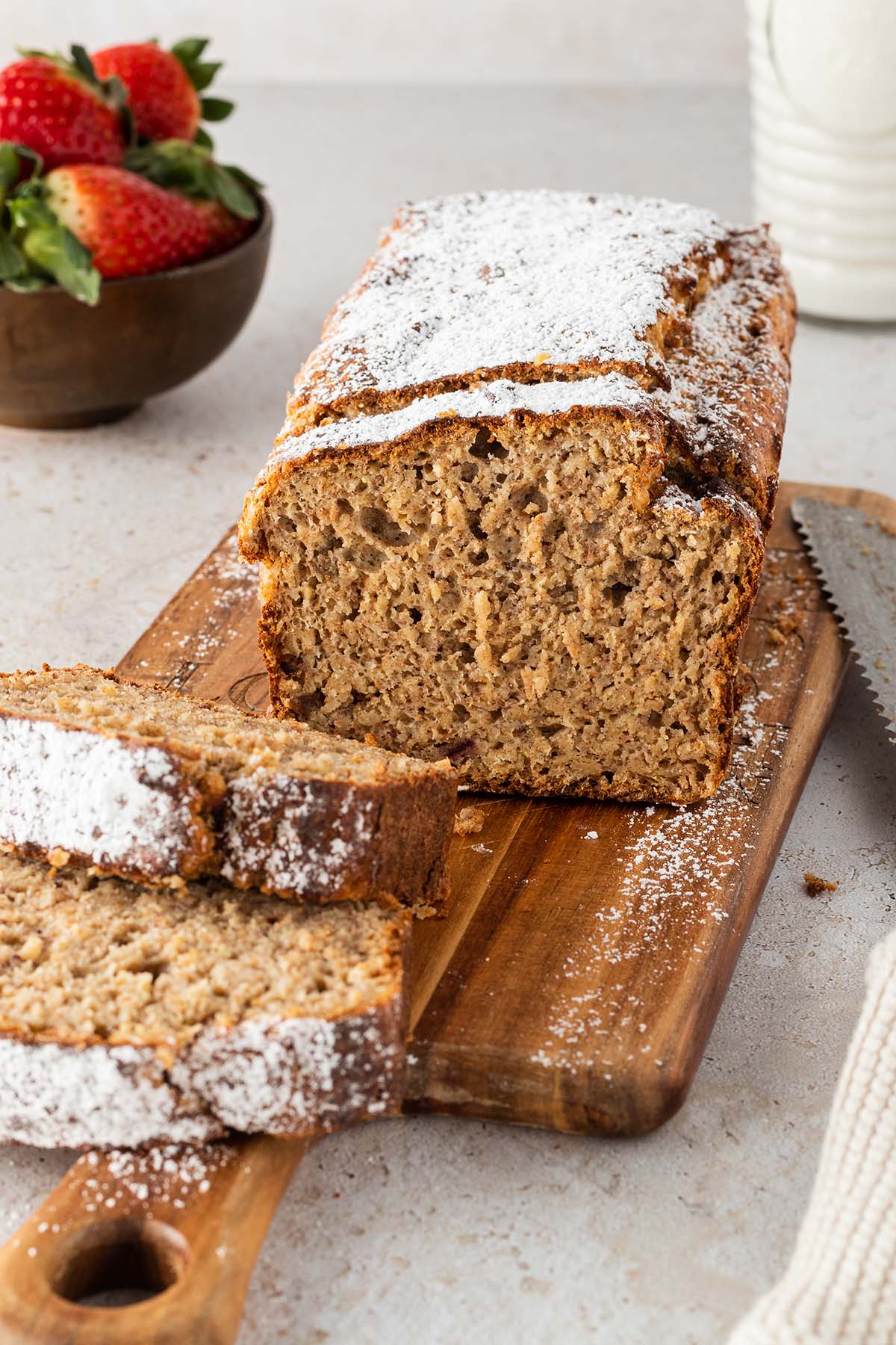Ein angeschnittenes Bananenbrot mit Puderzucker auf einem Holzbrett mit Deko im Hintergrund des Bildes.