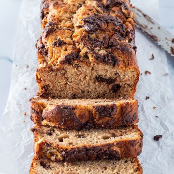 Ein angeschnittenes Bananenbrot mit Schokolade auf einem hellen Untergrund, mit einem Messer und einem Glas Milch im Hintergrund.