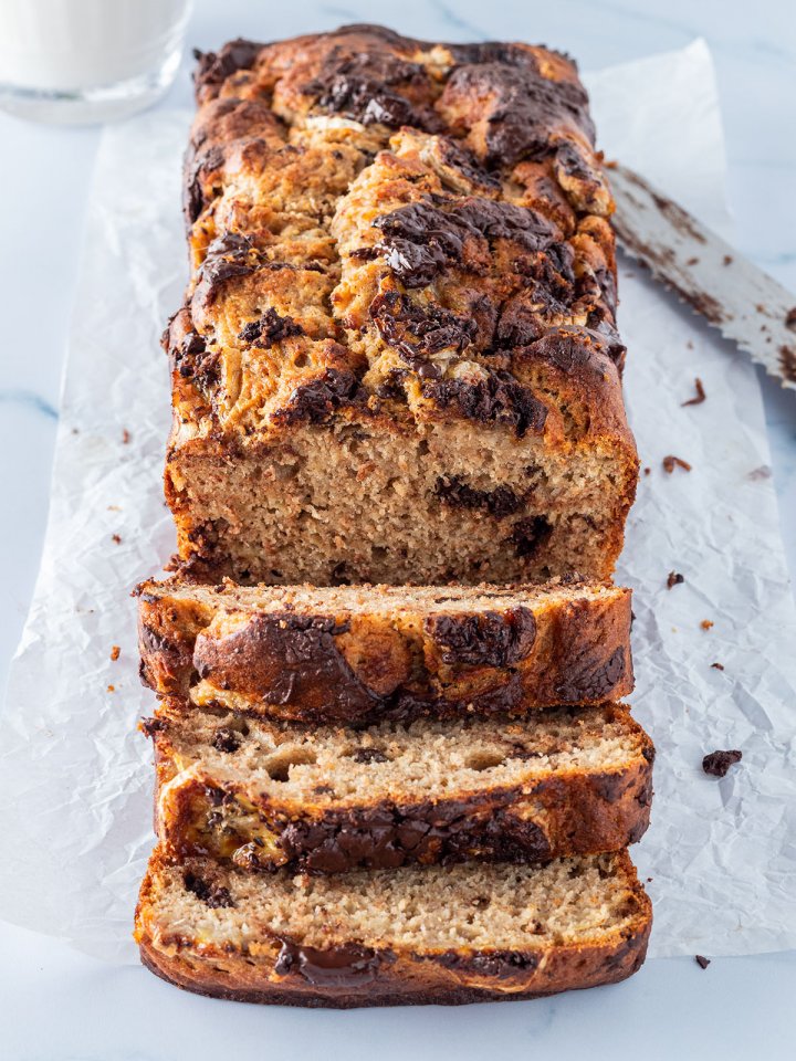 Ein angeschnittenes Bananenbrot mit Schokolade auf einem hellen Untergrund, mit einem Messer und einem Glas Milch im Hintergrund.