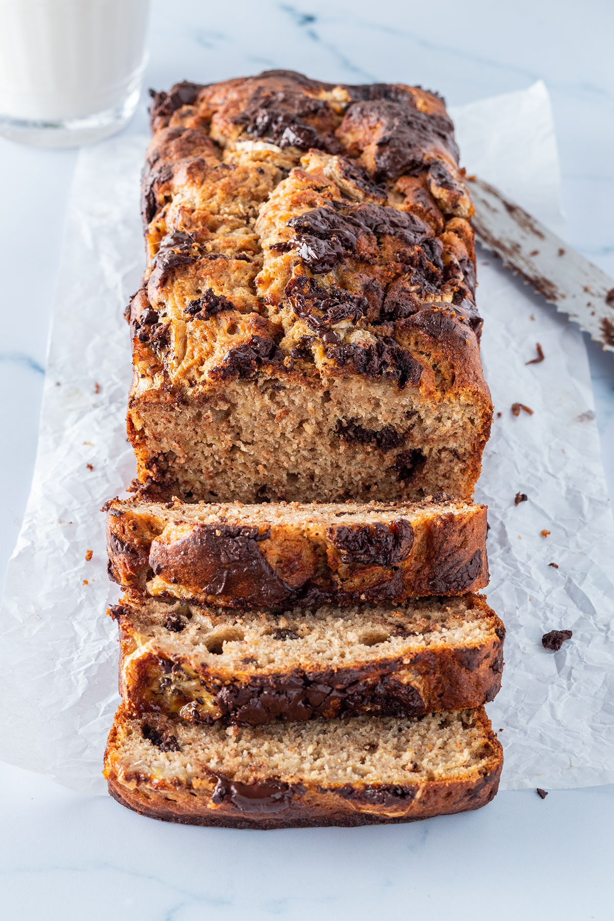 Ein angeschnittenes Bananenbrot mit Schokolade auf einem hellen Untergrund, mit einem Messer und einem Glas Milch im Hintergrund.