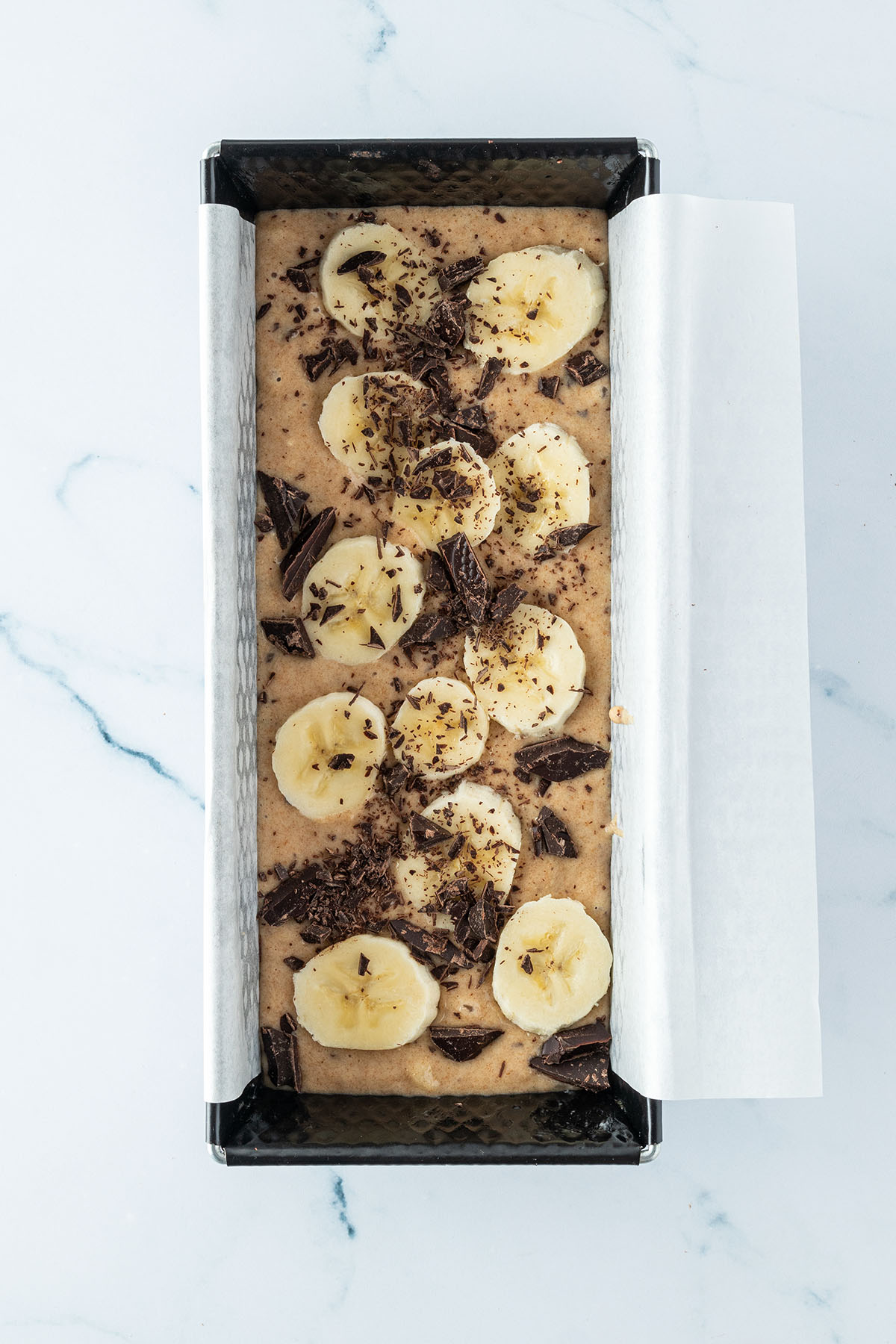 Teig f&uuml;r das Bananenbrot in einer Kastenform, mit Bananenscheiben und gehackter Schokolade auf der Oberfl&auml;che.