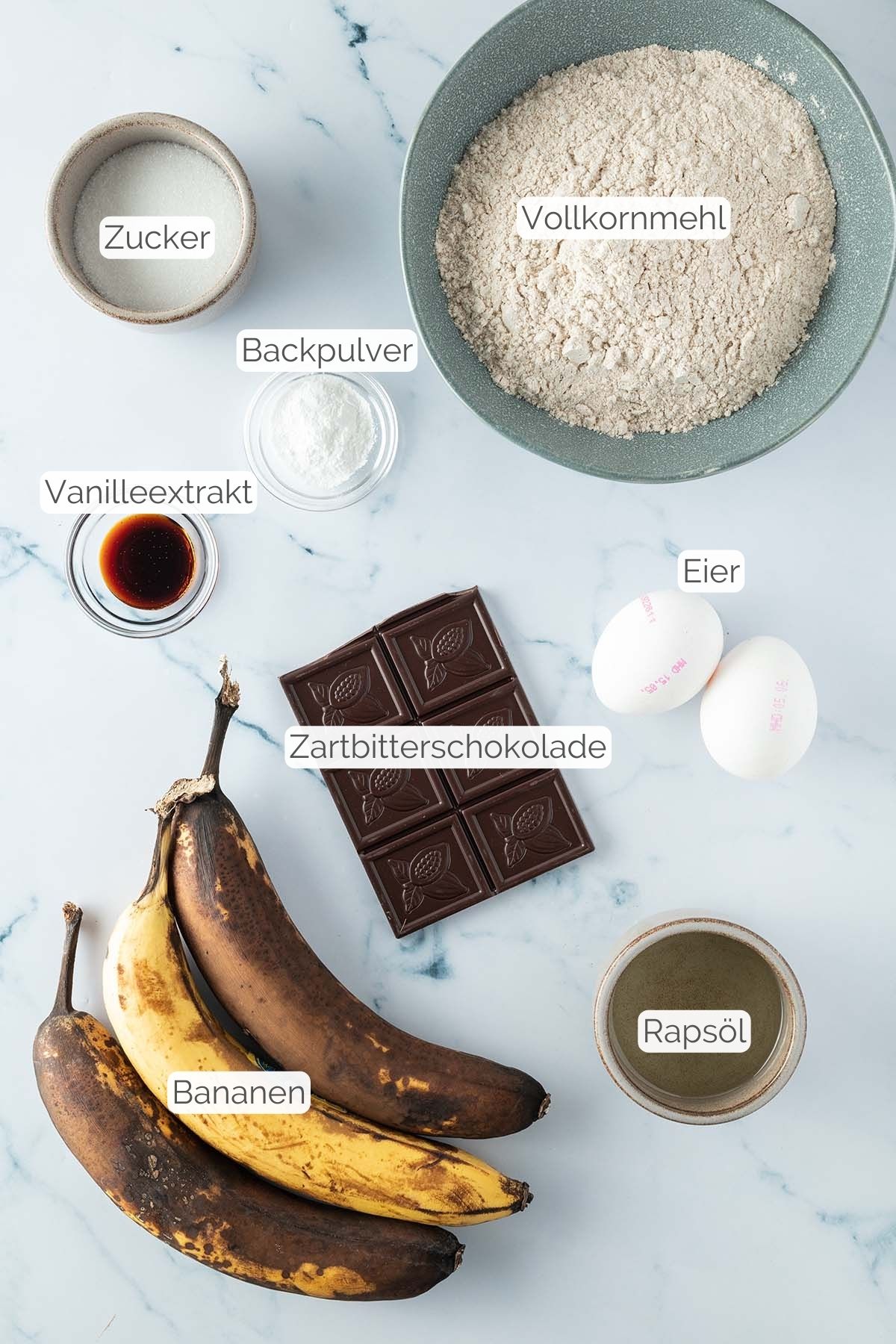 Foto aller Zutaten mit jeweiligen Textbeschriftungen, die f&uuml;r das Bananenbrot mit Schokost&uuml;ckchen ben&ouml;tigt werden.