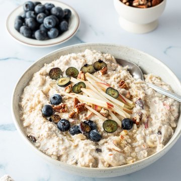 Bircher M&uuml;sli mit frischem Obst und N&uuml;ssen in einer Sch&uuml;ssel, frische Blaubeeren und N&uuml;sse im Hintergrund.