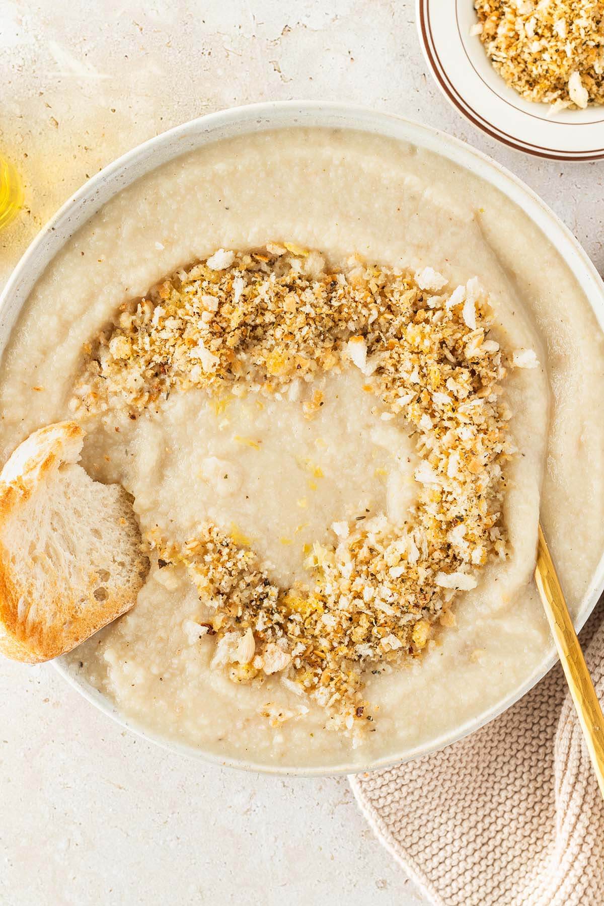 Teller mit Blumenkohlsuppe und einem Brotkr&uuml;mel-Topping in dem ein St&uuml;ck Brot getunkt wird, Deko im Bildrand.