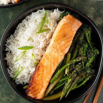 Bowl mit gebratenem Lachs, Reis und asiatischem Gem&uuml;se auf dunkelgr&uuml;nem Untergrund, Deko und zweite Sch&uuml;ssel im Bildrand.