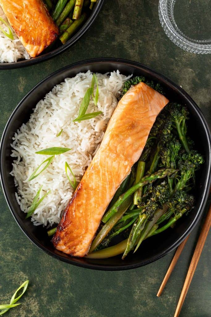 Bowl mit gebratenem Lachs, Reis und asiatischem Gem&uuml;se auf dunkelgr&uuml;nem Untergrund, Deko und zweite Sch&uuml;ssel im Bildrand.