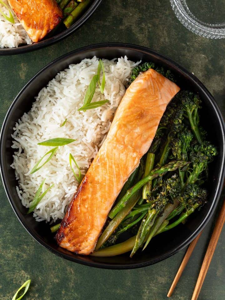 Bowl mit gebratenem Lachs, Reis und asiatischem Gem&uuml;se auf dunkelgr&uuml;nem Untergrund, Deko und zweite Sch&uuml;ssel im Bildrand.