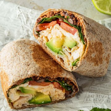 Halbierter Breakfast Burrito mit R&uuml;hrei, Bohnen, Avocado, Spinat und Tomaten F&uuml;llung auf einem St&uuml;ck Zeitungspapier. Deko im Hintergrund.