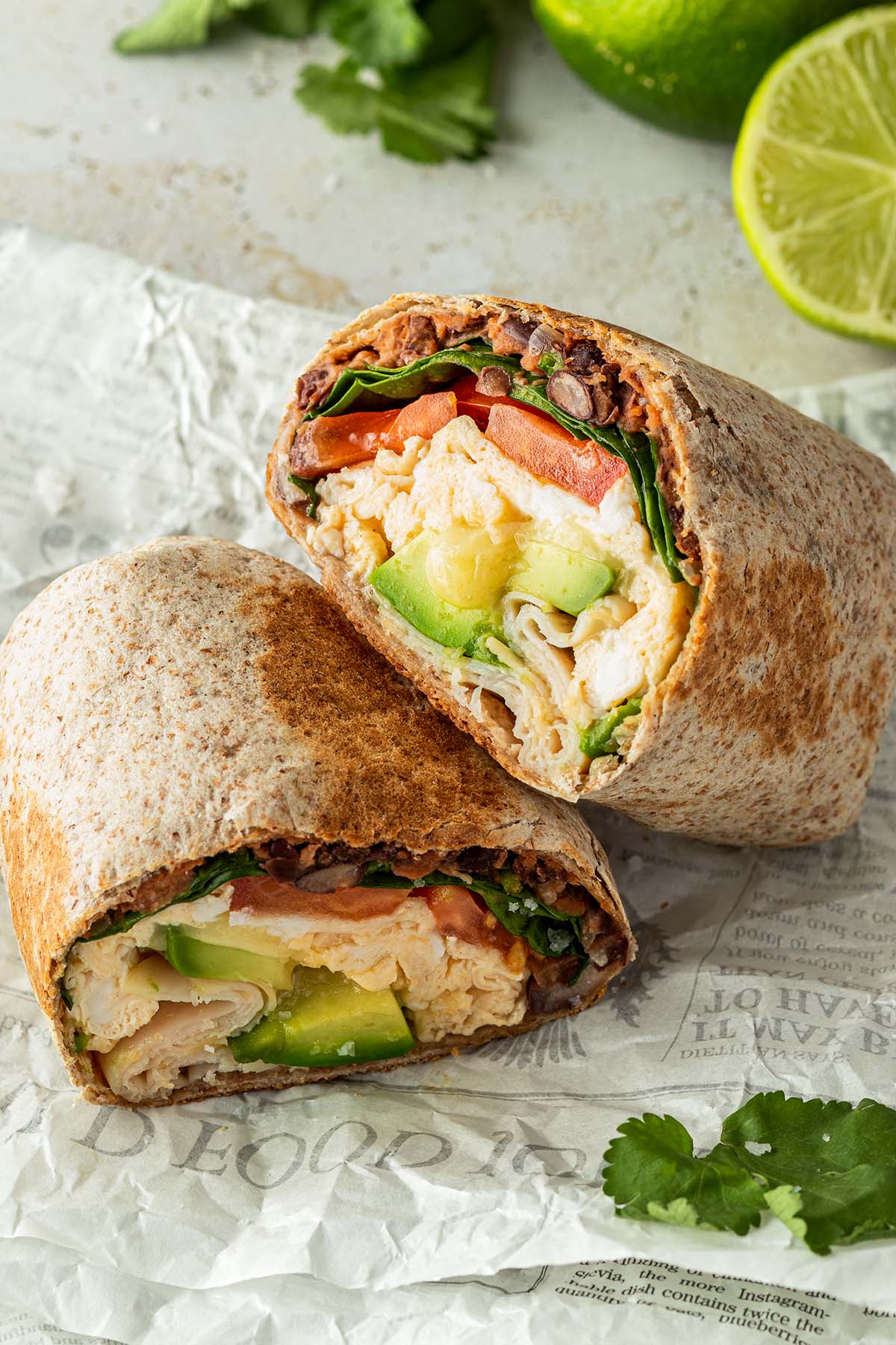 Halbierter Breakfast Burrito mit R&uuml;hrei, Bohnen, Avocado, Spinat und Tomaten F&uuml;llung auf einem St&uuml;ck Zeitungspapier. Deko im Hintergrund.