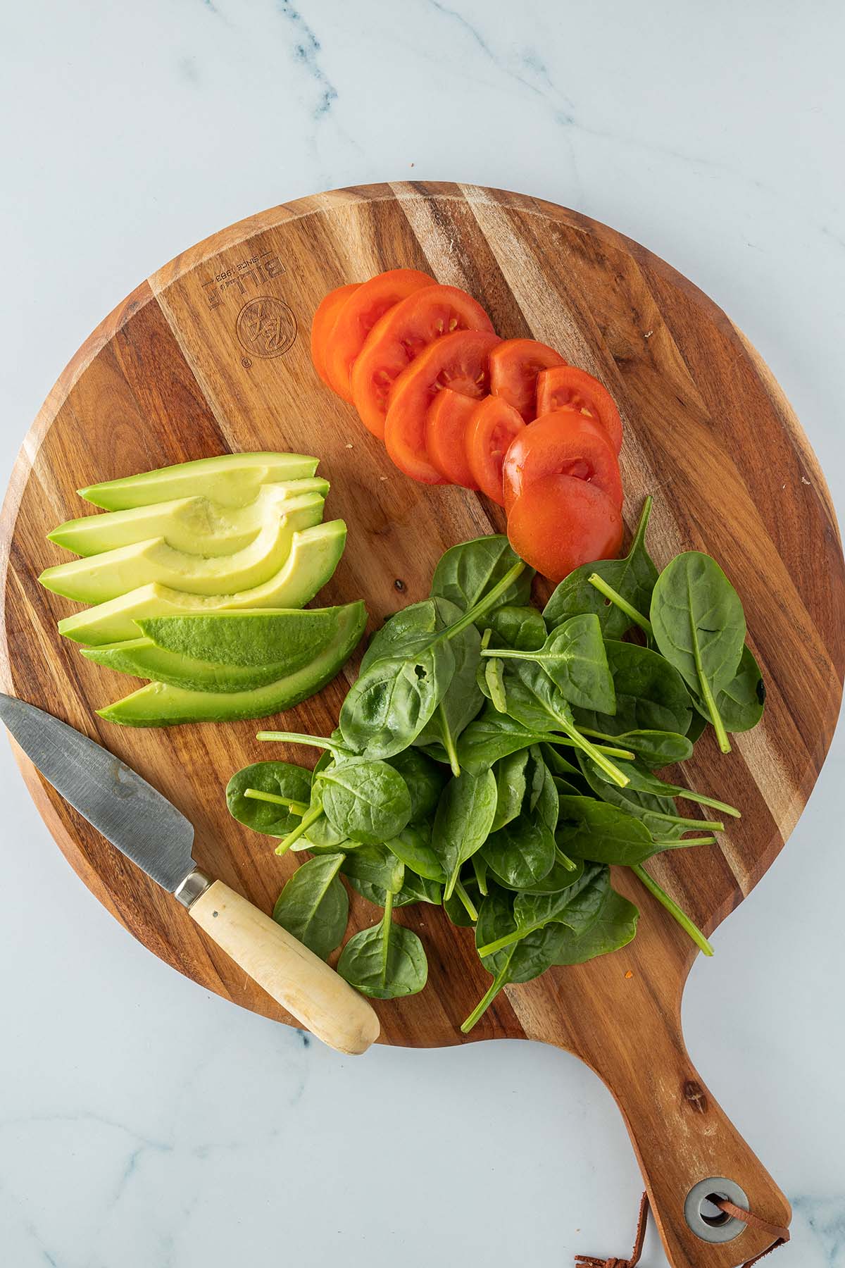 Ein Holzbrett mit einer in Scheiben geschnittenen Tomate, d&uuml;nne Avocadospalten und kleinen Spinatbl&auml;ttern.
