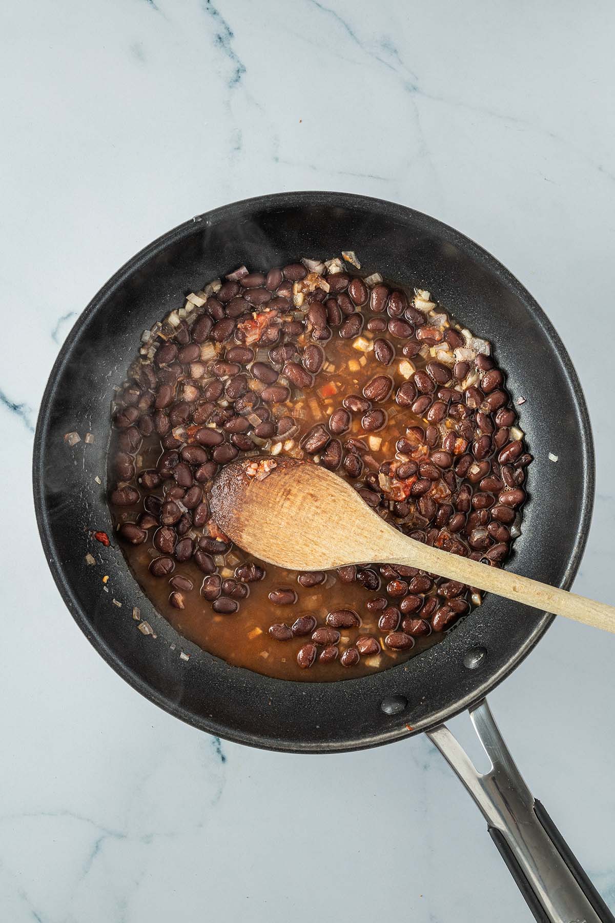 Schwarze Bohnen und kleingeschnittene Zwiebelst&uuml;cke in einer Pfanne mit etwas Fl&uuml;ssigkeit.