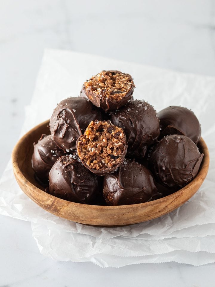 Mehrere ganze und halbierte, mit Schokolade &uuml;berzogene Brownie Energy Balls in einer kleinen Holzsch&uuml;ssel.