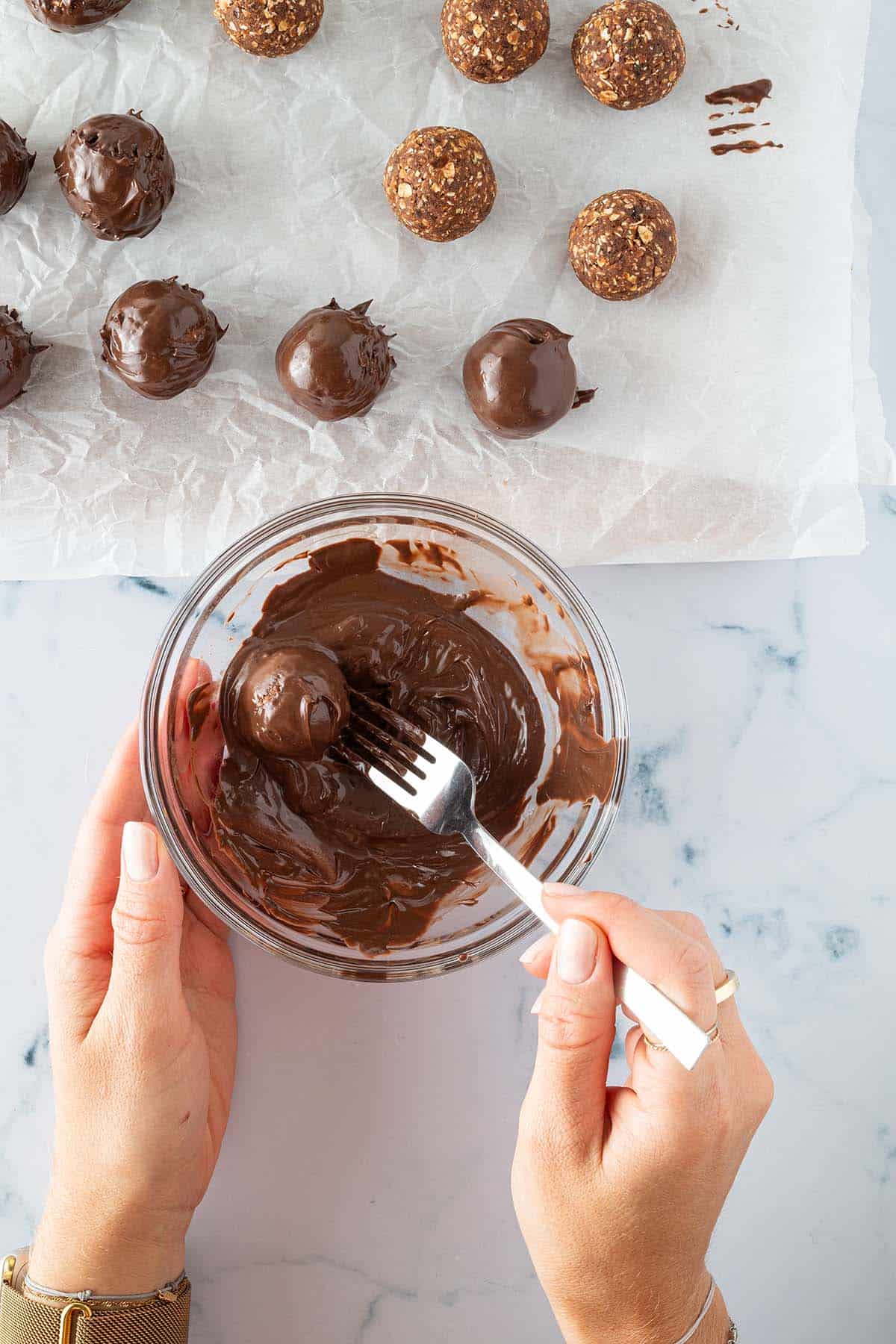 Die zu Kugel geformten Dattel-Schoko-B&auml;llchen werden in einer Sch&uuml;ssel geschmolzener Schokolade gew&auml;lzt. Daneben ein Backpapier mit Schokolade &uuml;berzogener Brownie Balls.