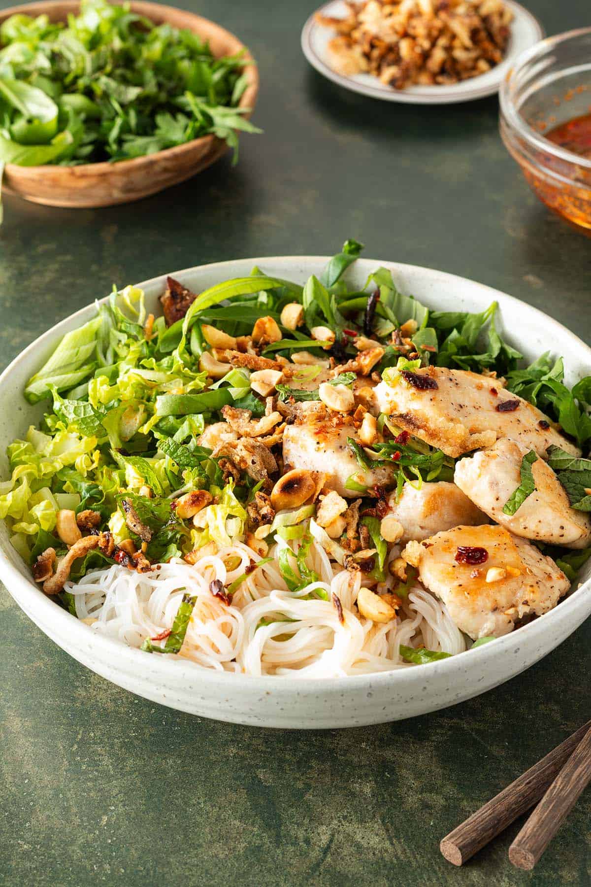 Vietnamesischer Reisnudelsalat mit H&auml;hnchen, Erdn&uuml;ssen, frischen Kr&auml;utern und R&ouml;stzwiebeln in einer Sch&uuml;ssel, im Hintergrund kleine Sch&uuml;sseln mit Zutaten.