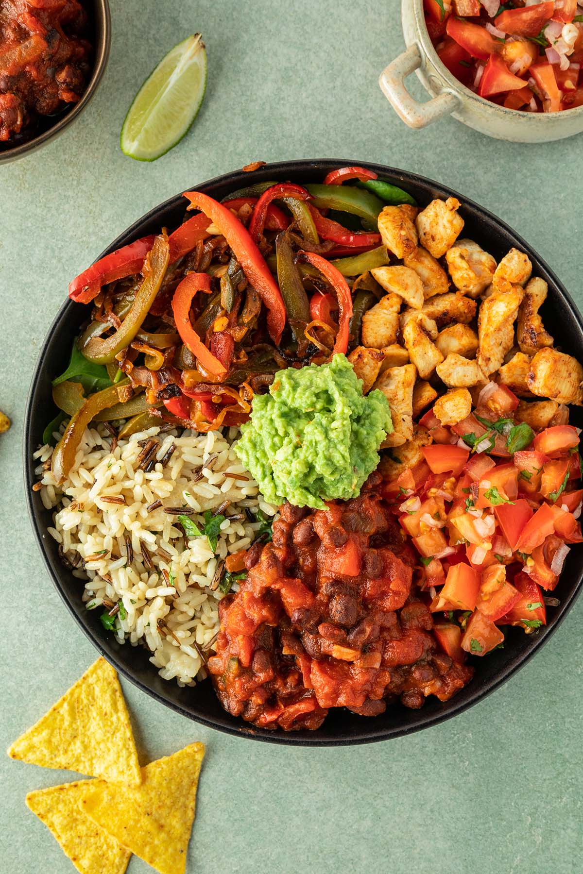 Selbstgemachte Burrito Bowl in einem tiefen Teller mit verscheidenen Komponenten, die nebeneinander angeordnet sind. Daneben eine Sch&uuml;ssel mit Salsa, eine Limetten-Spalte und Nachos.