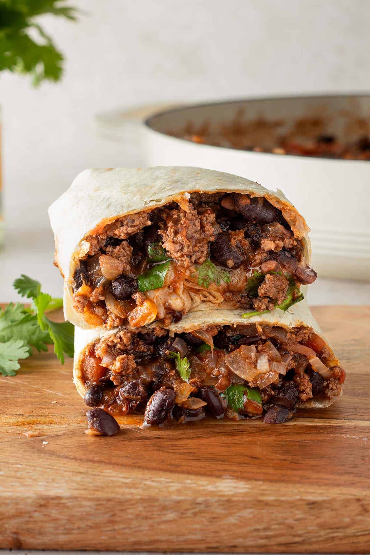 Ein in der Mitte halbierter Burrito gef&uuml;llt mit einer Hackfleisch und Bohnen Pfanne auf einem Holzbrett, im Hintergrund eine Pfanne mit Chili con Carne F&uuml;llung und frische Kr&auml;uter.