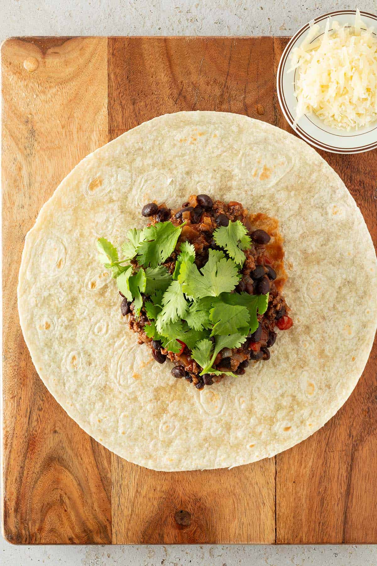 Ein Tortilla auf einem Brett mit der Hackfleisch F&uuml;llung und frischem Koriander in der Mitte. Daneben geriebener K&auml;se auf einem kleinen Teller.