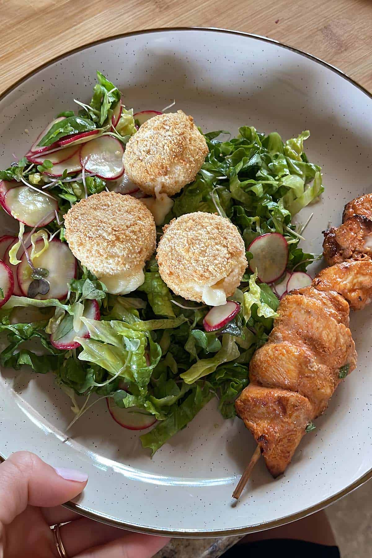 Drei kleine, im Airfryer gebackene Camemberts auf gemischten Salat auf einem Teller mit einem H&auml;hnchenspie&szlig;.