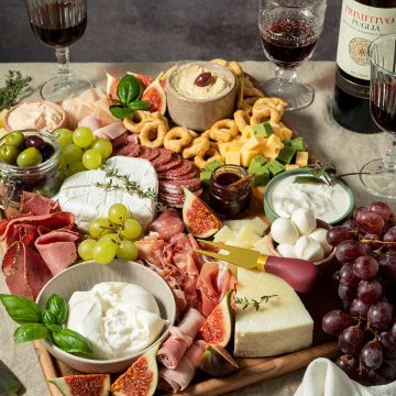 Gro&szlig;e Charcuterie-Platte mit K&auml;se, Salami, Schinken, Feigen, Trauben, Crackern, Dips und Rotwein auf einem gedeckten Tisch.