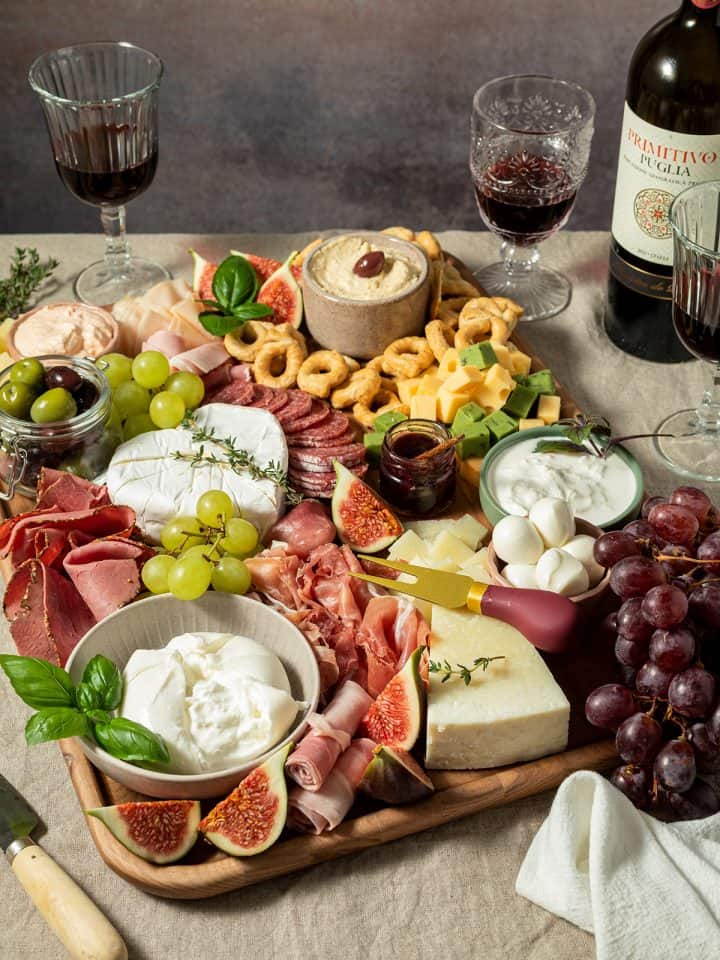 Gro&szlig;e Charcuterie-Platte mit K&auml;se, Salami, Schinken, Feigen, Trauben, Crackern, Dips und Rotwein auf einem gedeckten Tisch.
