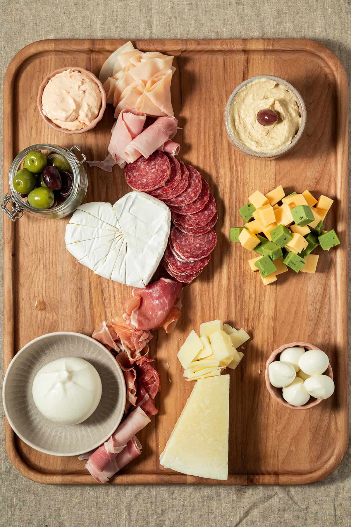 Charcuterie-Platte in der Aufbauphase mit K&auml;se, Wurst, Trauben, Feigen, Dips und Oliven auf einem Holzbrett.